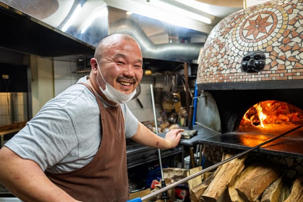 Image of smiling pizza maker standing in front of his stone oven making a fresh pizza